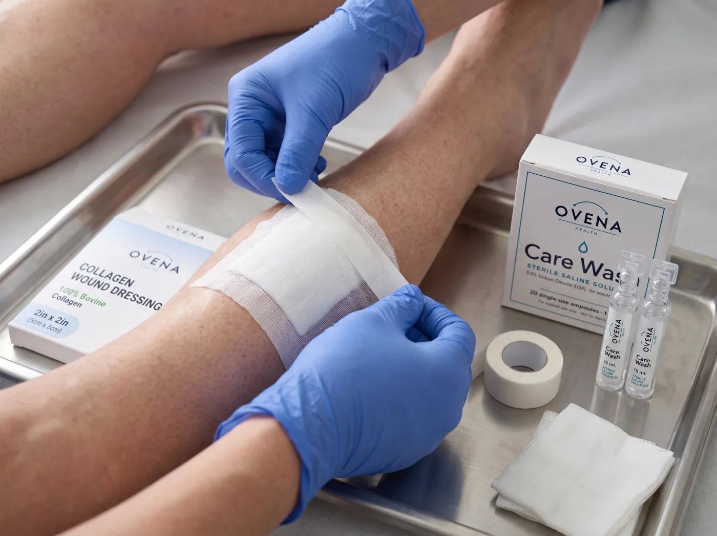 Gloved hands smooth paper tape over a gauze secondary dressing on a lower leg, with the Ovena Collagen Wound Dressing box, the Care Wash carton and two ampoules on the tray beside it.