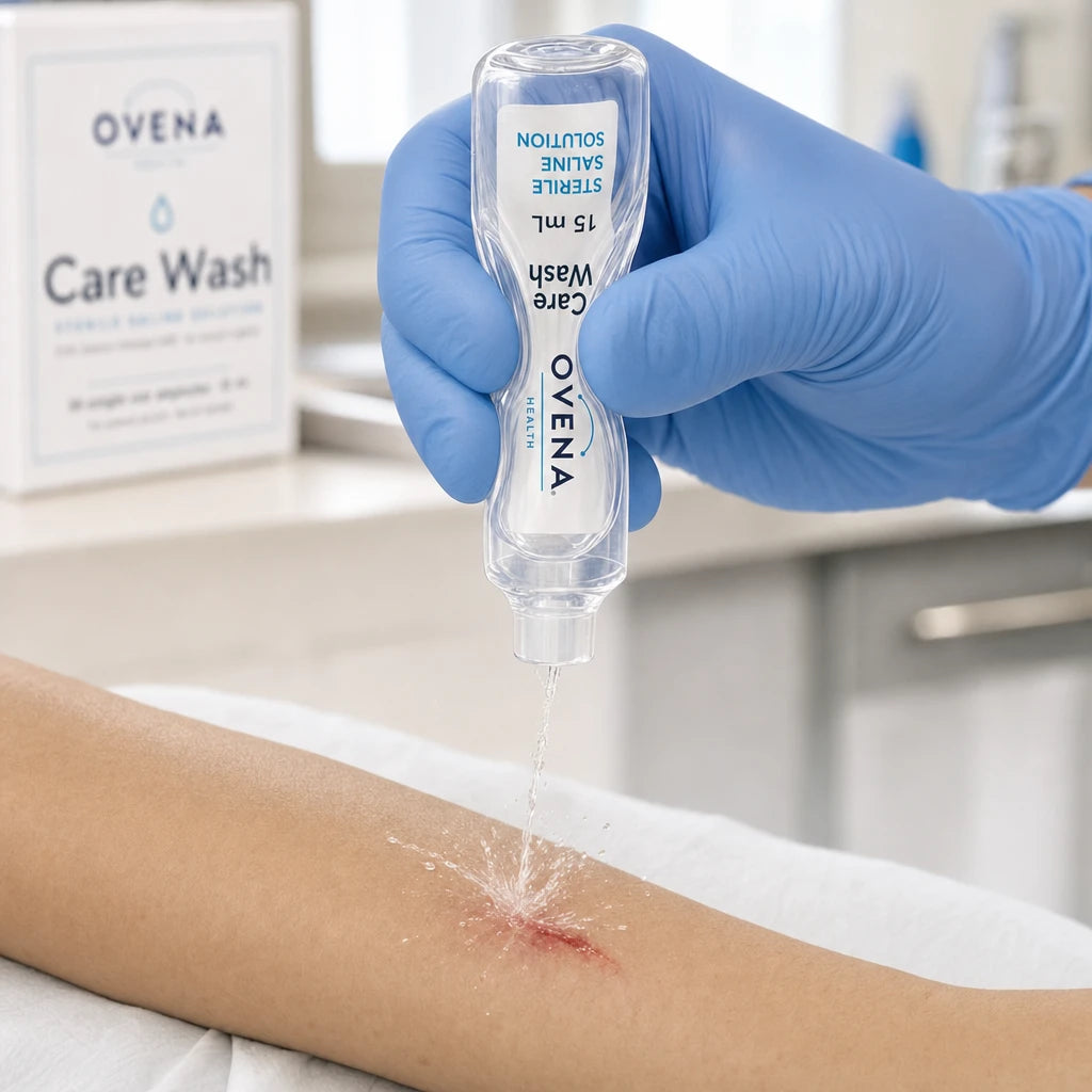 A gloved hand squeezing an opened Care Wash ampoule so a jet of sterile saline strikes a graze on the forearm.