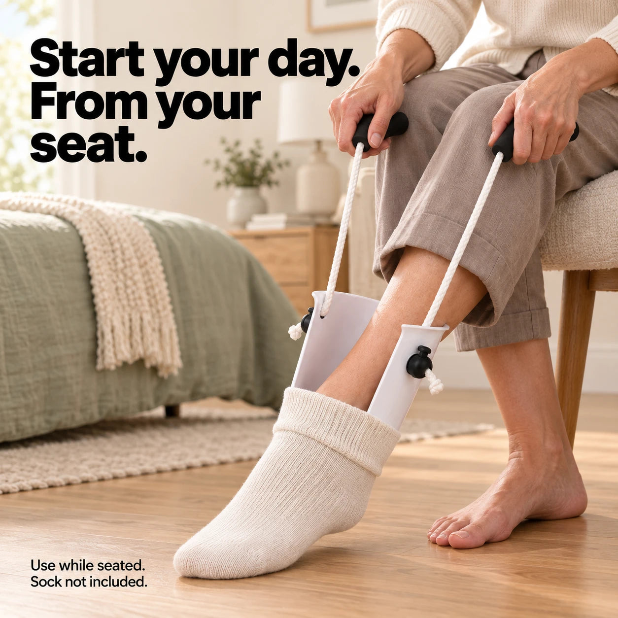 Seated on the edge of a bed, both cord handles pulled, the white sock aid shell drawing a cream sock up over the foot.