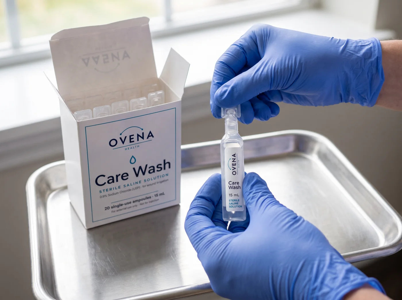 Gloved hands snap the flat tab off an Ovena Care Wash ampoule over a stainless tray, with the open Care Wash carton and its sealed ampoules beside them.