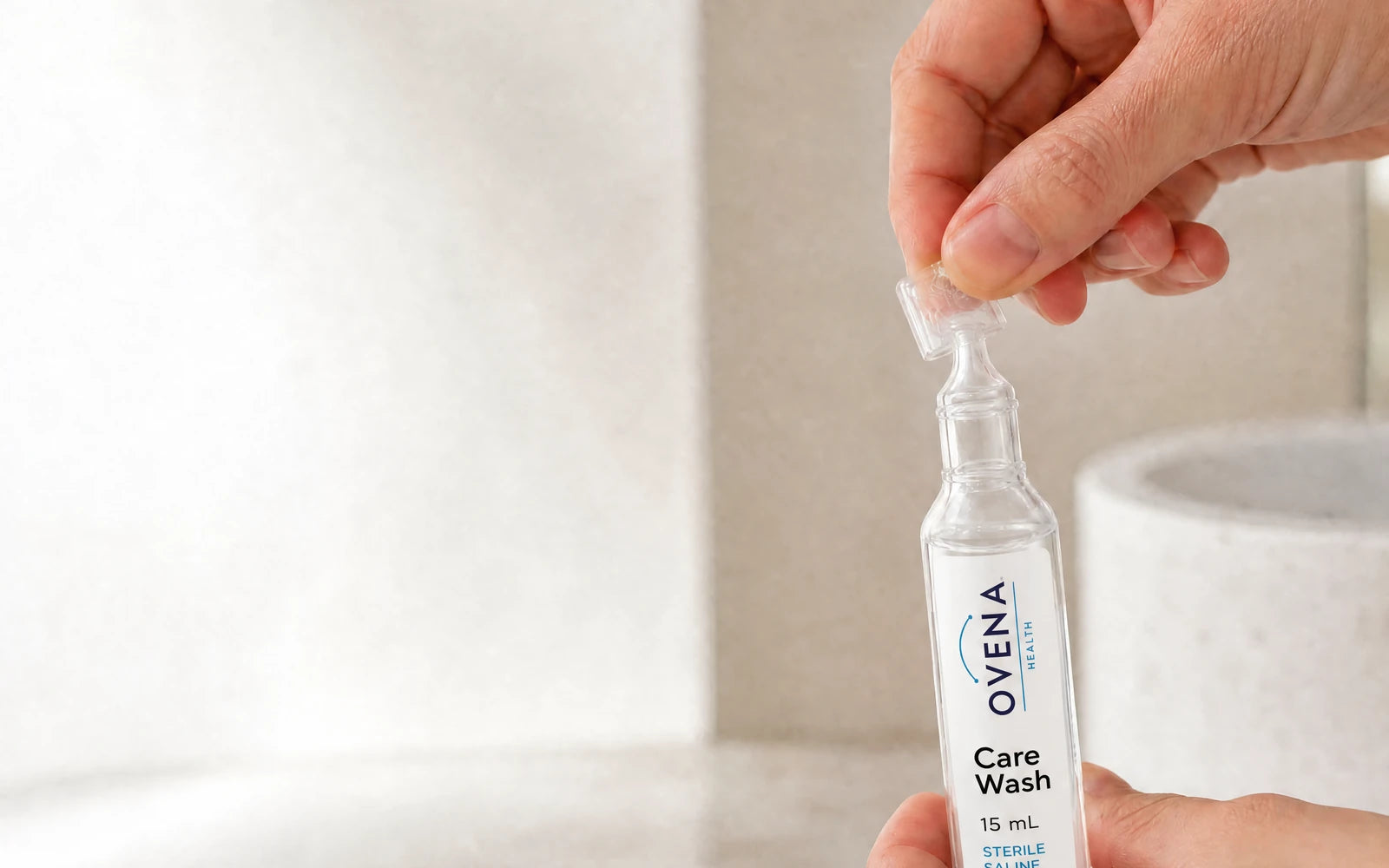 Hands twisting the tab off an Ovena Care Wash saline ampoule at a bathroom counter